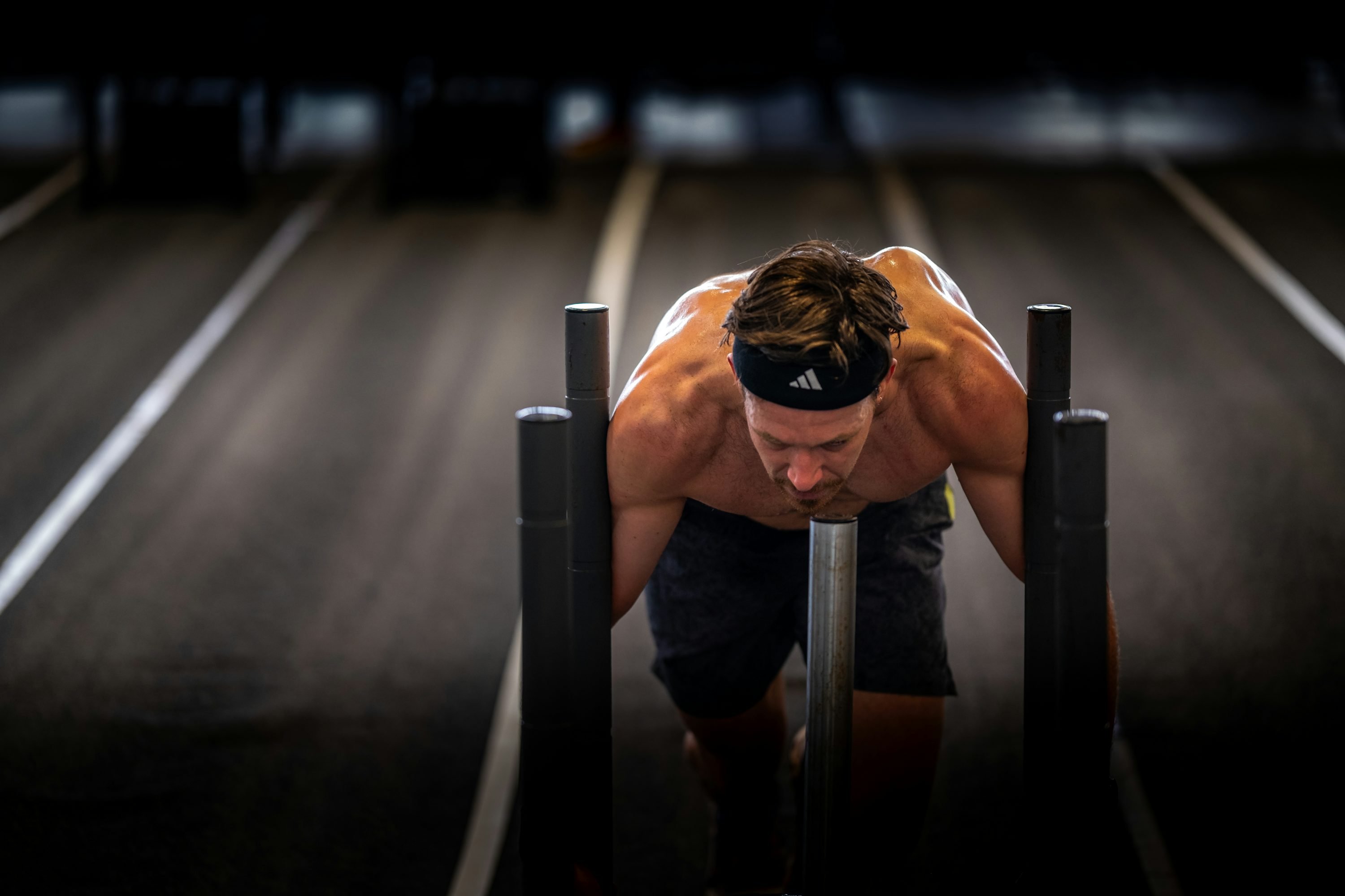 Athlète concentré en sprint de traînée, têtière et buste appuyé sur un traîneau à pousser, dans une zone sombre de gymnase. Athlète concentré en sprint de traînée, têtière et buste appuyé sur un traîneau à pousser, dans une zone sombre de gymnase.