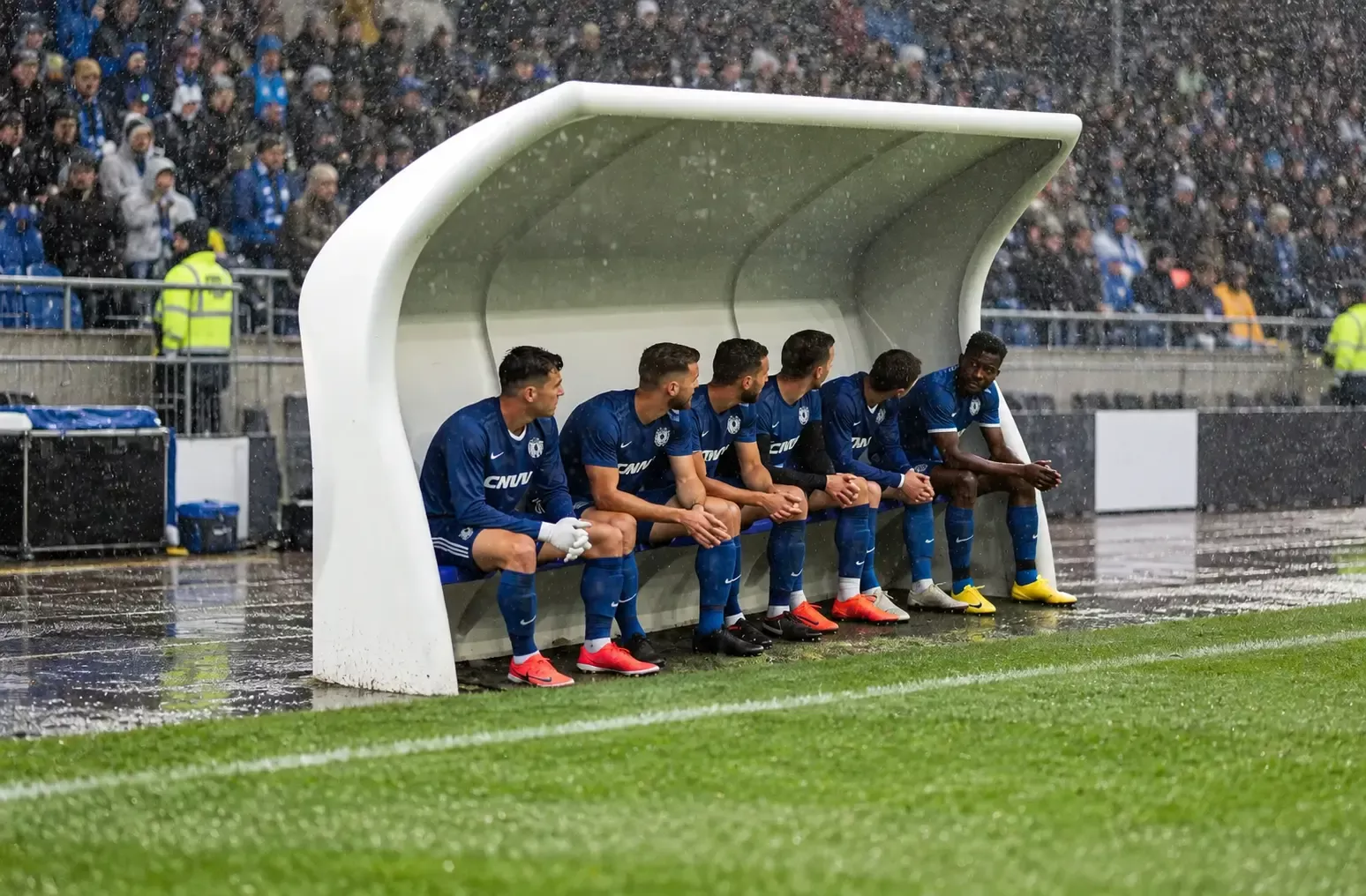 Équipe de football en ère sous la pluie dans le dugout blanc, joueurs en maillots bleus assis sur le banc.
