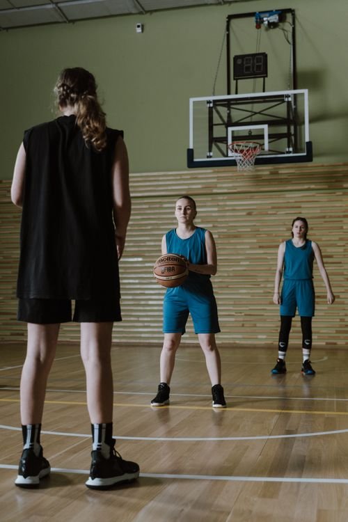 Trois joueurs de basket en salle, une femme au centre avec le ballon, spectateur au premier plan.